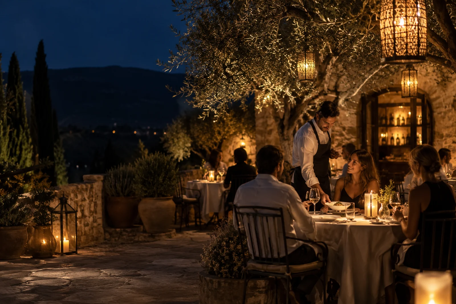 Terrasse de restaurant de nuit, tables dressées et lanternes ambrées sous les oliviers.